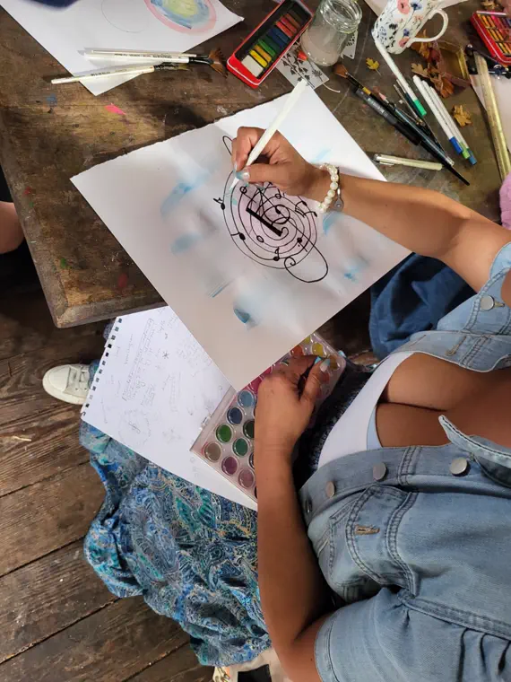 Close-up of a hand drawing a black ink circular mandala on paper, with pencils and a watercolour tin alongside.