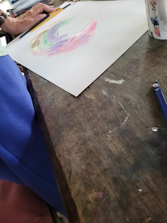 Close-up of a hand holding a pencil beside pink, green and yellow pastel strokes on paper on a wooden table.