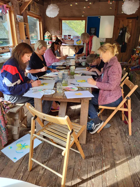 People drawing and painting at a long table covered in artwork inside The Larch Barn workshop.
