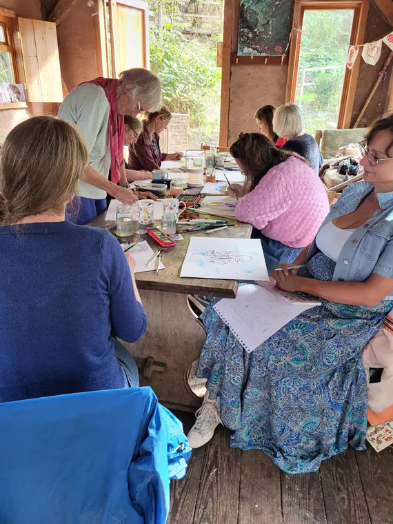 Women drawing and painting at a long table in a timber barn while the facilitator leans over to help.